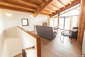 A spacious room with a high ceiling, wooden beams, a grey sofa, a small table with a TV, and a window with curtains.