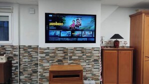 A living room with a flat-screen TV mounted on the wall displaying a show, a wooden cabinet, and a stone-patterned backsplash. - Loft Los Olivos in Algar (Algar)