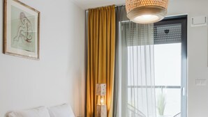 Habitación de hotel con sofá verde, almohadas blancas, una obra de arte enmarcada en la pared, una ventana con cortinas amarillas y vistas al exterior.