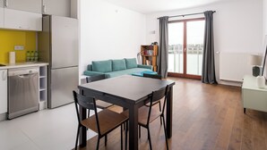 Een moderne keuken met witte kasten, een roestvrijstalen koelkast en een vaatwasser. Er is een eethoek met een houten tafel en stoelen. Op de achtergrond staat een blauwgroene bank, en er is een raam met gordijnen te zien.