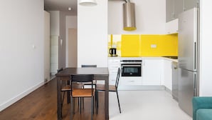 Een moderne keuken met witte kasten, een geel accentmuur, roestvrijstalen apparatuur en een houten eettafel met stoelen.