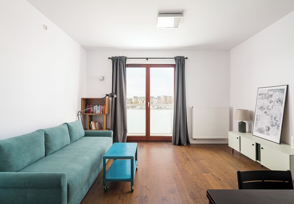 Un salon moderne avec un canapé turquoise, une petite table bleue, une bibliothèque, un radiateur et une vue à travers une porte vitrée.