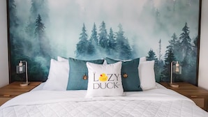 Un lit recouvert d’une courtepointe blanche matelassée, un oreiller orné d’un motif de canard et une fresque murale sur le thème de la forêt.
