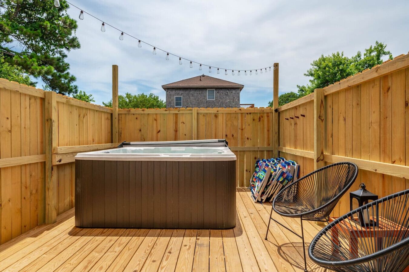 Une terrasse en bois avec un jacuzzi, deux chaises en fil de fer noir et une guirlande lumineuse.