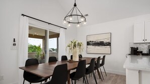 A modern dining area with a wooden table and black chairs, a hanging light fixture, and a view of greenery outside. - Serenity House-Water Views, Pet Friendly & Hot Tub (Kill Devil Hills)
