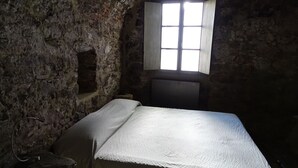 A stone-walled room with a single bed, a small window, and a stone bench.
