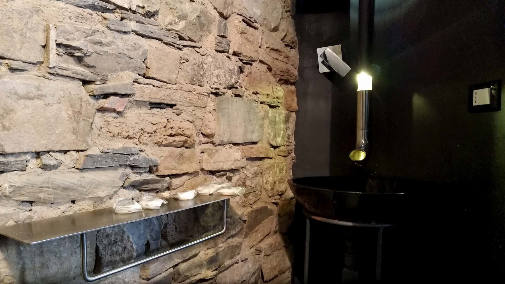 Una pared de piedra con un estante metálico que sostiene velas, un aplique de pared y un taburete negro.