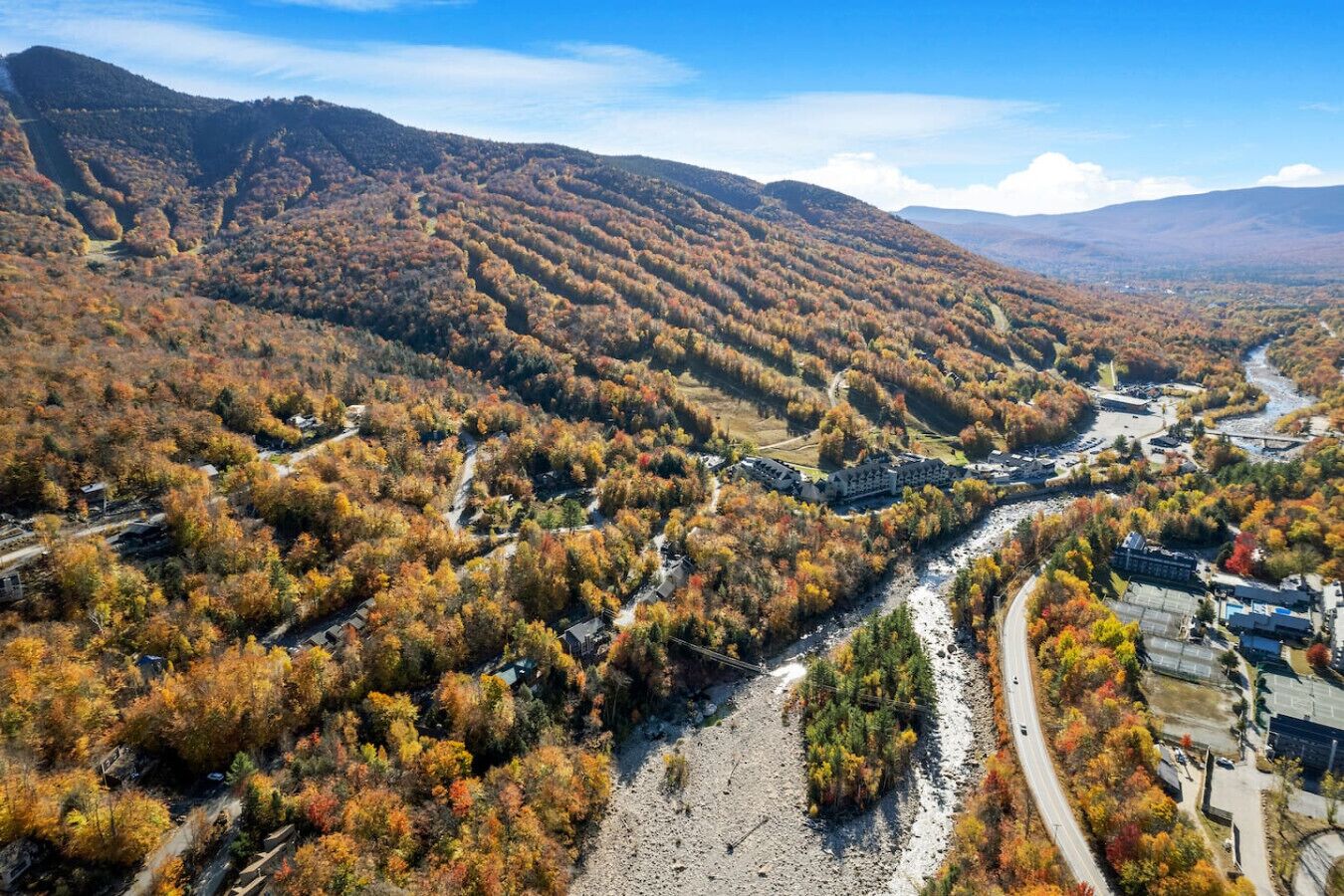 Luftaufnahme eines Tals mit einem Fluss, umgeben von herbstlich gefärbten Bäumen und einer Bergkulisse.