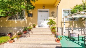 Un edificio con fachada amarilla, una puerta blanca y escalones que conducen a la entrada. Hay plantas en macetas en los escalones y un banco a la derecha.