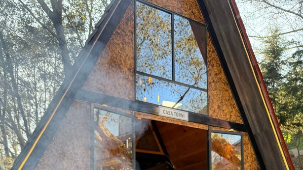 Una rustica baita di legno con una griglia di fronte, fumo che sale e un cartello con su scritto “CASA TORNI”.