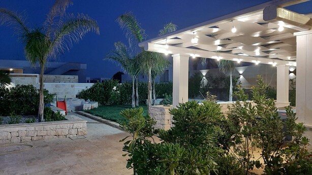 A covered outdoor area with a stone wall, palm trees, and a pergola with lights. - Villa Romolo with swimming pool 150 mt from the Caribbean beach of San Pietro (San Pietro In Bevagna)