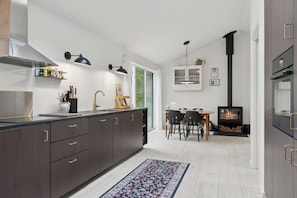 A modern kitchen with dark cabinetry, a central island, pendant lighting, a dining area with a table and chairs, and a wood-burning stove.