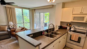 Una cocina compacta con una isla integrada, un fregadero y un horno. Hay una ventana con persianas, un ventilador de techo y una lámpara en la mesita de noche.
