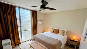 Una habitación de hotel con cama, ventilador de techo, banco, mesita de noche con lámpara y ventana con cortinas.