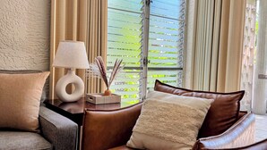 Una sala de estar moderna con un sofá y sillón de cuero, una mesita de noche con l ámpara y objetos decorativos, y ventanas amplias con persianas.