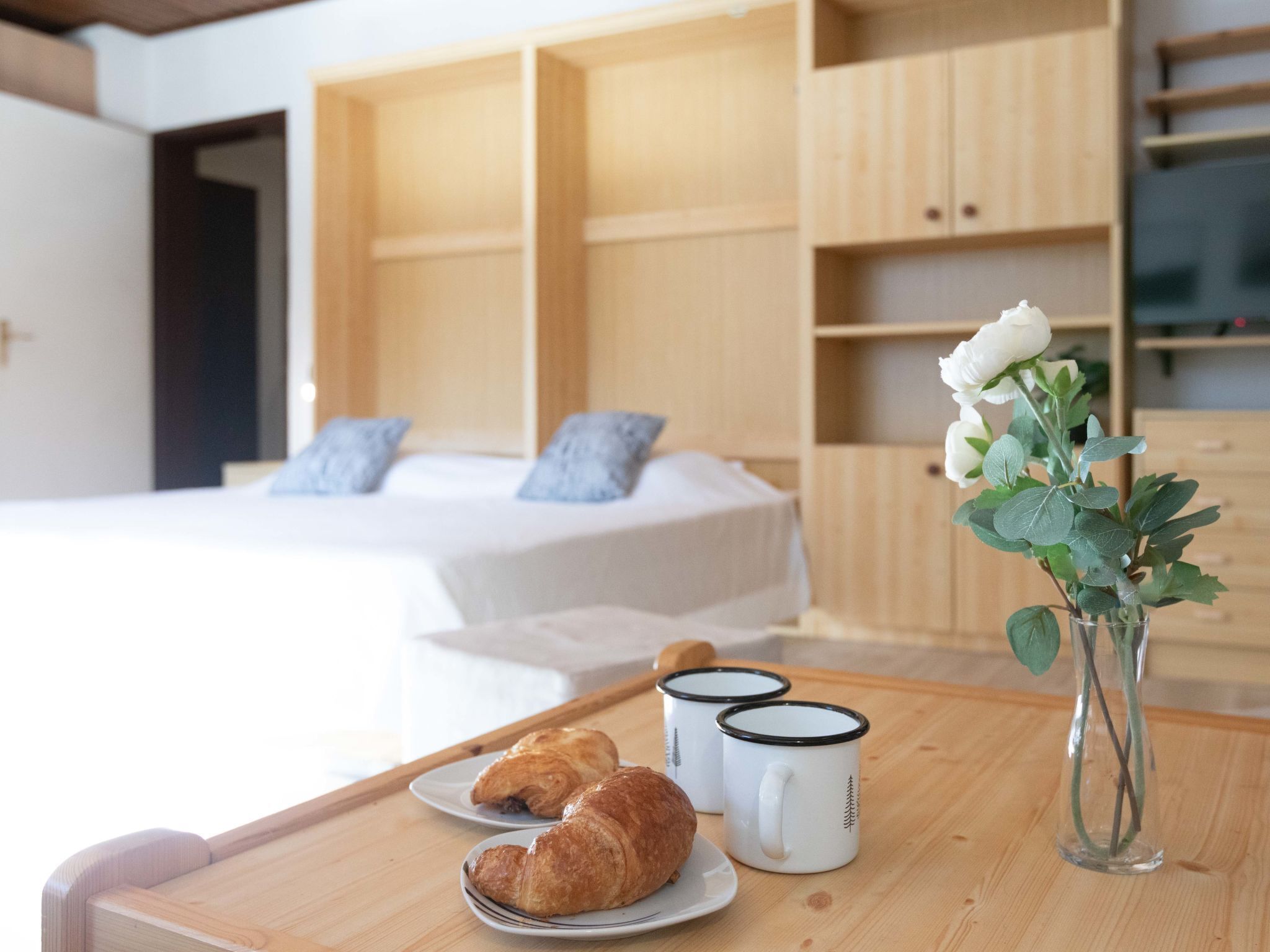 A bedroom with a bed, a wooden nightstand, a vase with a white flower, and two mugs on the nightstand.