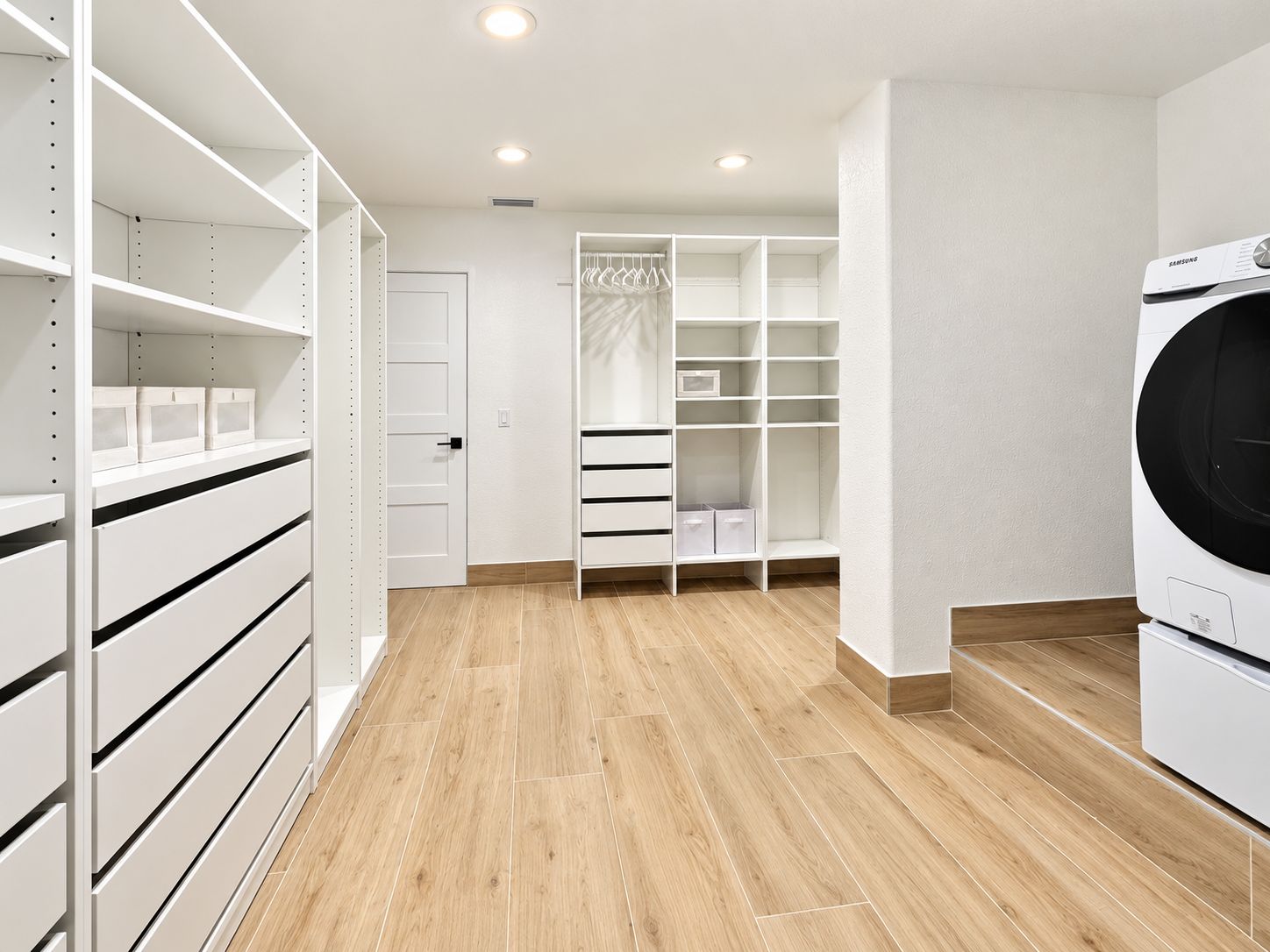 A laundry room with a washing machine, a white closet with shelves and drawers, and a wooden floor.