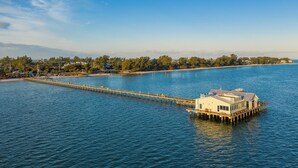 A waterfront property with a long pier extending into the water, a large house on stilts, and a beach in the background.