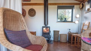 A cozy room with a wicker chair, a small table, a fireplace, and a window with a view of trees. - The Hut, Walberswick (Walberswick)