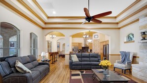 A spacious living room with a dark gray sofa, a matching armchair, and a coffee table with a vase of yellow flowers. A ceiling fan is visible, and the room has wooden beams and a stone fireplace. - Luxury Ranch in Florence-Party House for Families 5 1/2 bedrooms,5bath,Sleeps 20 (Florence)