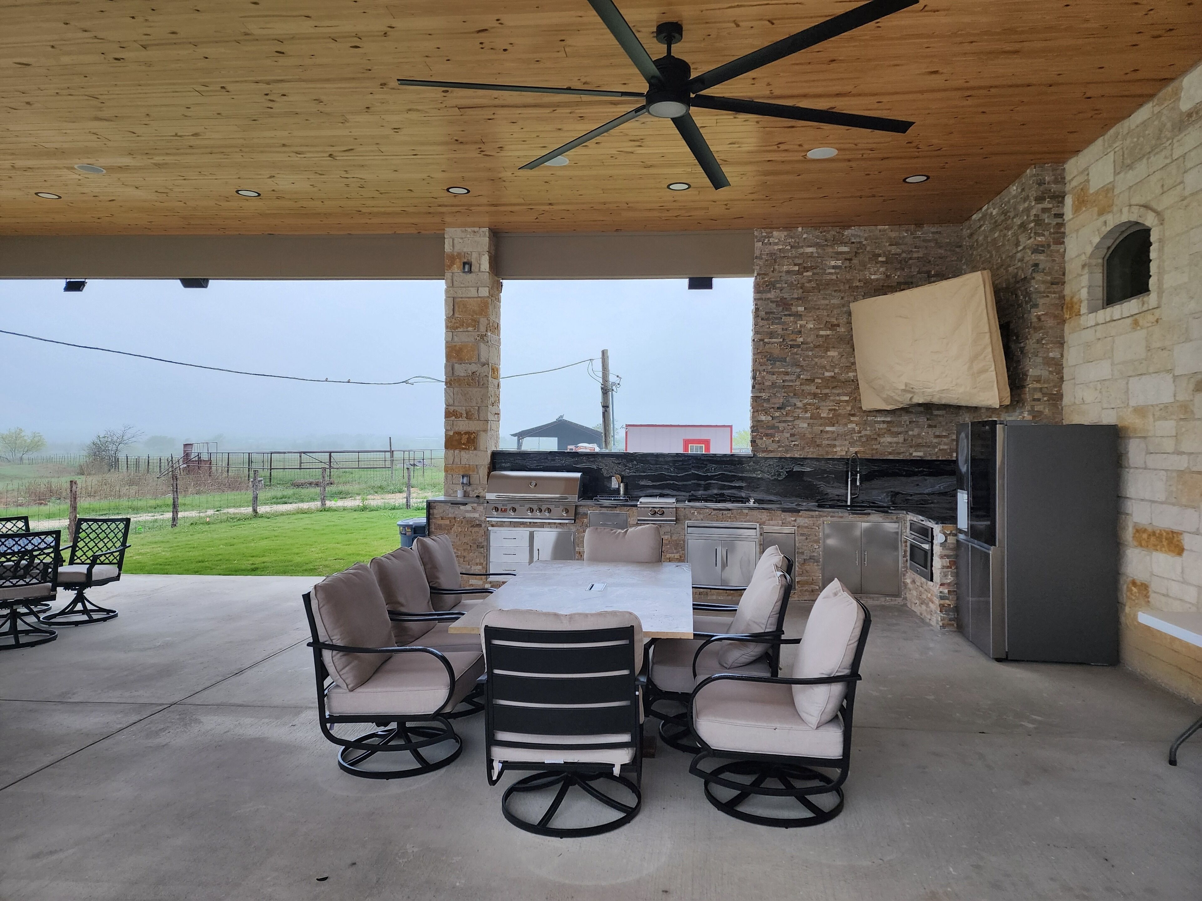 An outdoor patio with a wooden ceiling, stone pillars, a grill, and a refrigerator.