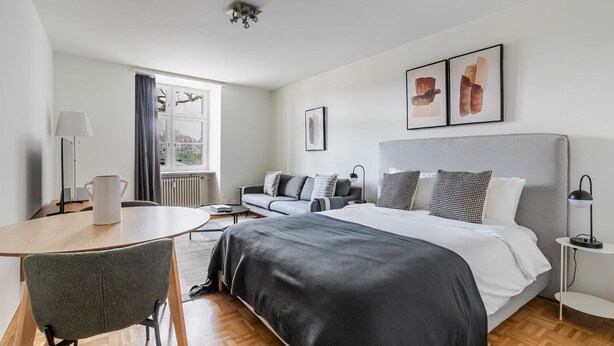 A modern hotel room with a large bed, a sofa, a dining table, and a chair. There is a window with curtains, a radiator, and a ceiling light. - Blueground | ID97, Basel-Vorstaedte, Washer-Dryer (Basel)
