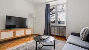 A modern living room with a flat-screen TV, wooden cabinet, round coffee table, grey sofa, and a view of the city through a window. - Blueground | ID97, Basel-Vorstaedte, Washer-Dryer (Basel)