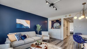 Uma sala de estar moderna com um sofá cinzento, uma mesa de centro redonda e uma cadeira azul. Há uma área de cozinha com armários brancos e uma mesa de jantar com cadeiras azuis.