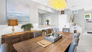 A dining area with a wooden table, chairs, a chandelier, a large painting, and a staircase.