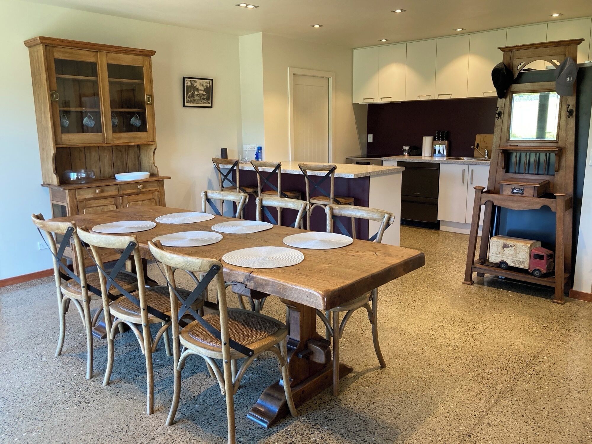 A dining area with a wooden table set for six, surrounded by wicker chairs. A wooden cabinet with glass doors is to the left, and a kitchen area is visible in the background.