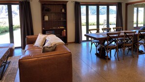 A spacious living area with a brown leather sofa, a wooden dining table with chairs, and a bookshelf. Large windows provide a view of the outdoors.