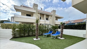 A modern building with a flat roof, a balcony, and a well-maintained lawn with two lounge chairs. - Appartamento Forte 3 - Carraro Immobiliare (Lido di Jesolo)