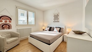 A bedroom with a bed, bedside tables, a chair, a window, and a radiator. - Appartamento Forte 3 - Carraro Immobiliare (Lido di Jesolo)