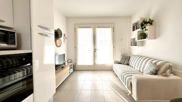 A compact living space with a kitchenette, a sofa, and a door leading to an outdoor area. - Appartamento Forte 3 - Carraro Immobiliare (Lido di Jesolo)