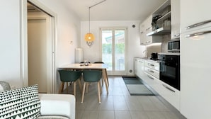 A modern kitchen with white cabinets, a dining area with a table and chairs, and a pendant light. - Appartamento Forte 3 - Carraro Immobiliare (Lido di Jesolo)