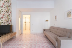 A living room with a TV on a wooden stand, a beige sofa, patterned carpet, and a door leading to another room.