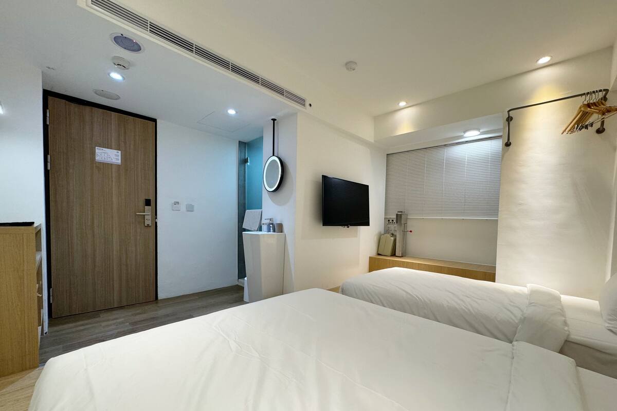 A hotel room with two beds, a TV, a mirror, a vanity with a sink, and a window with blinds.