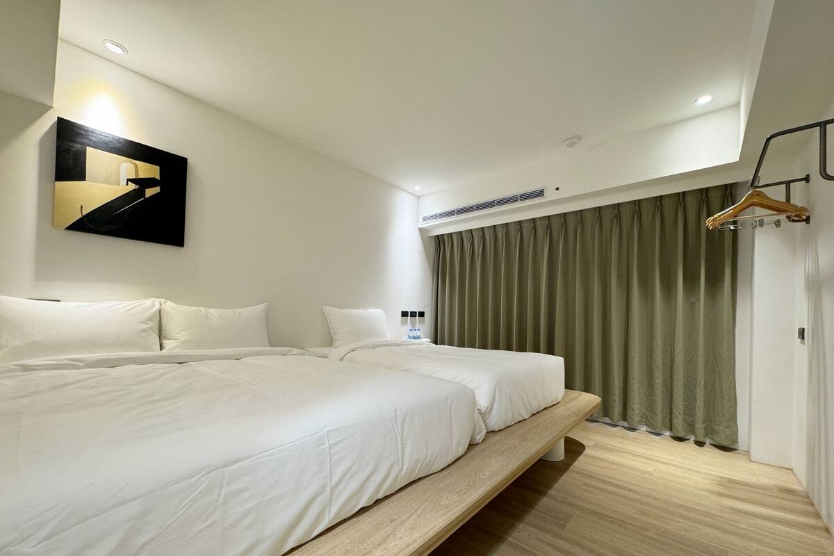 A modern hotel room with a large bed, a wooden bench, a wall-mounted light, and a window with curtains.