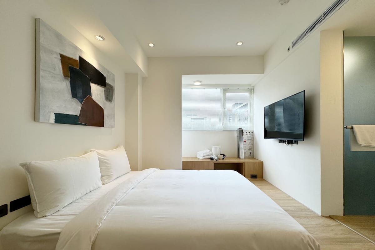 A modern hotel room with a large bed, a flat-screen TV mounted on the wall, a desk with a chair, and a window with blinds.
