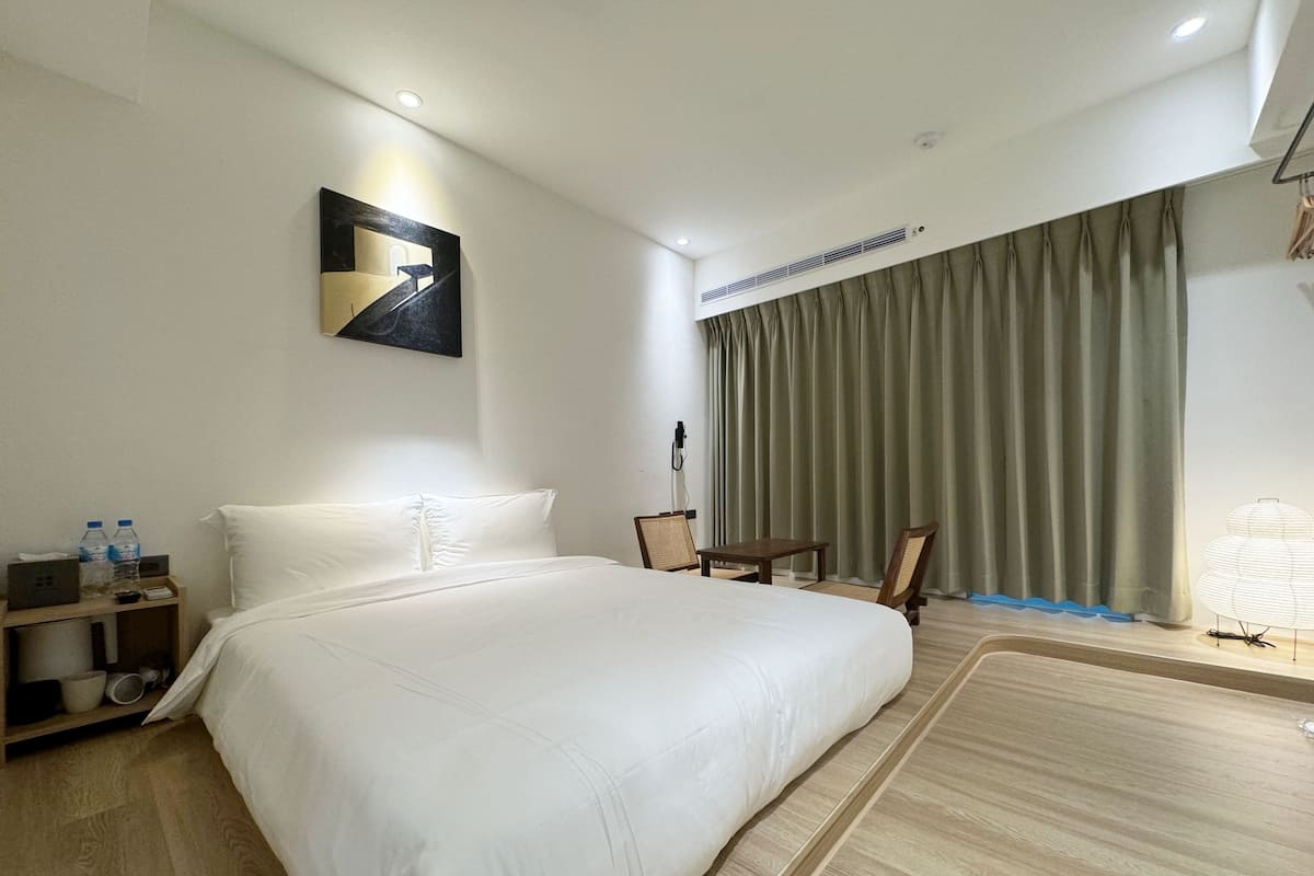 A hotel room with a large bed, a small table, a chair, a bedside table with a lamp, a painting on the wall, and a window with curtains.