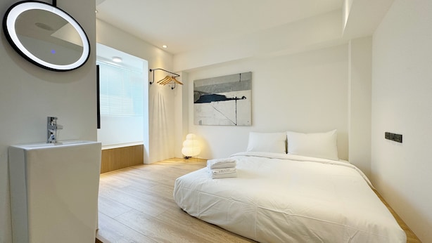 A modern bedroom with a large bed, a bedside table, a wall-mounted mirror, and a window with blinds. - A Day A Room (Taichung)