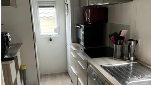 Cocina compacta con lavavajillas, estufa y pileta. Hay una ventana con persianas, un cesto de basura y una pequeña mesada con utensilios de cocina.