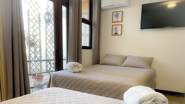 A hotel room with two beds, a large window with curtains, a flat-screen TV mounted on the wall, and a potted plant on the windowsill.