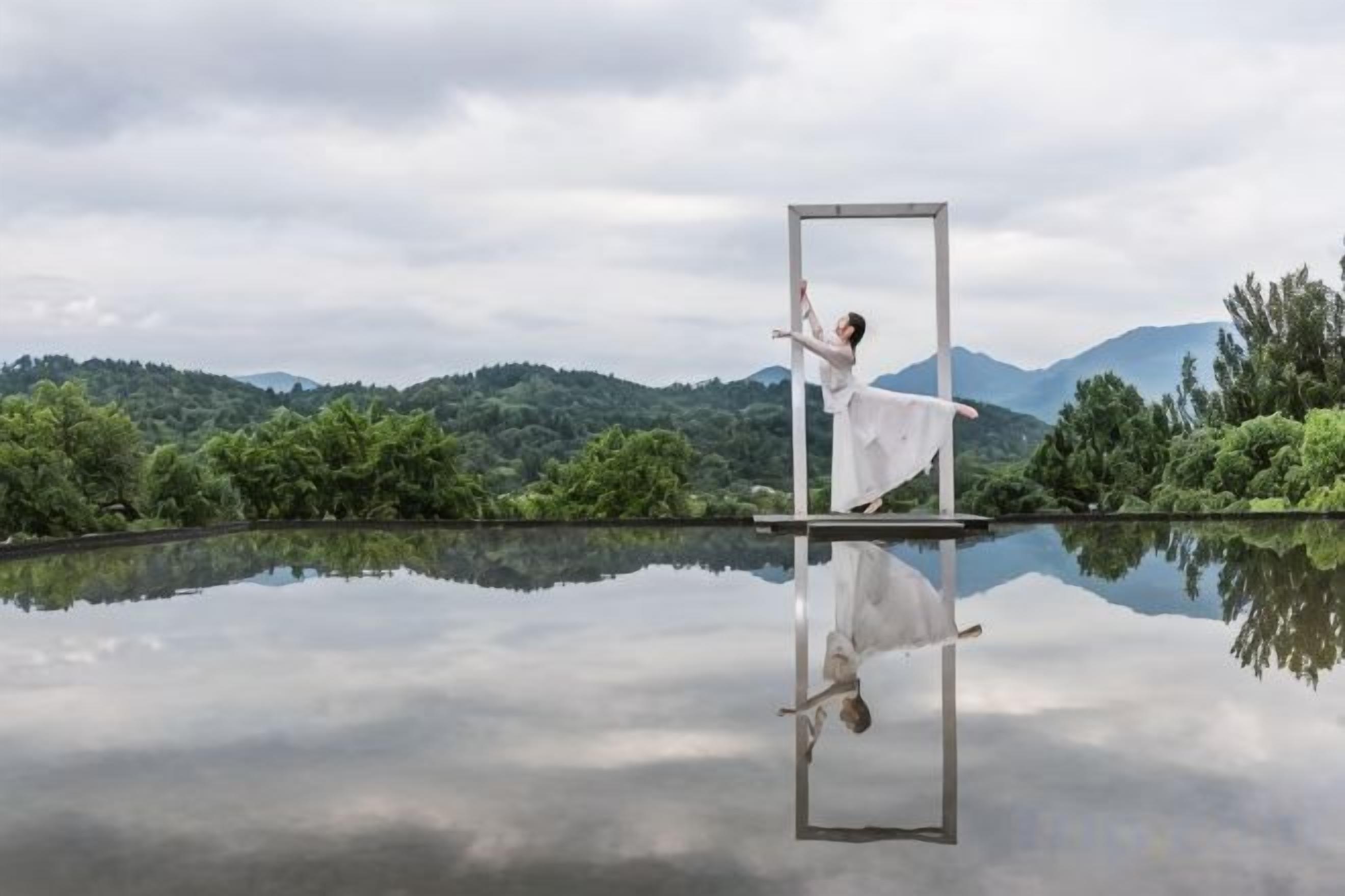 Una persona bailando frente a una superficie reflectante con un paisaje montañoso al fondo.