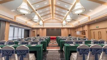 Sala de conferência com fileiras de mesas e cadeiras, um palco e teto alto com iluminação embutida.