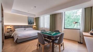 A hotel room with a bed, a desk with a lamp, a dining table with chairs, and a large window with curtains.