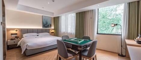 Habitación de hotel con cama, escritorio con lámpara, mesa de comedor con sillas y un ventanal con cortinas.
