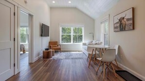 Une pièce spacieuse avec un plafond haut, un plancher en bois, une grande fenêtre, un canapé, une table à manger avec des chaises et un téléviseur fixé au mur.
