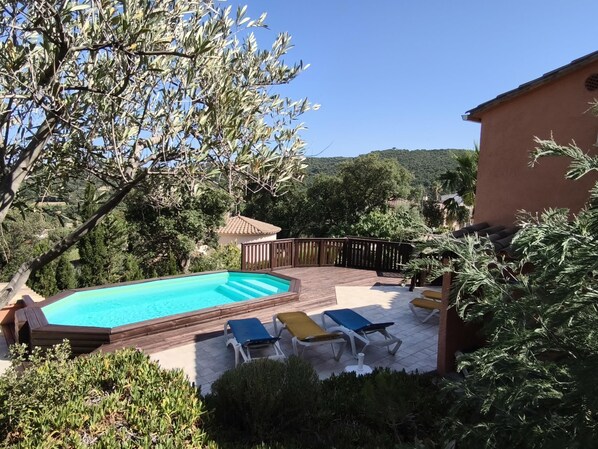 A swimming pool with a wooden deck, surrounded by greenery and a view of hills.