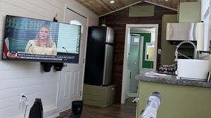 A cozy room with a wooden ceiling, a television displaying a news briefing, a refrigerator, and a kitchen area with a sink and stove.